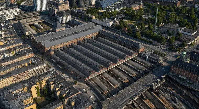 Aerial from the southwest: the great hall's long grey roof lies at the upper left with rooflight bands and corner turrets, the six glazing-striped barrel vaults run down to the right and end in semicircular glazed gables with red arches, and beyond them the tracks drop into a cutting under a road bridge. Vesterbro fills the lower left, Tivoli the upper right.