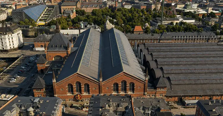 Low aerial from the west: the sunlit twin brick gable of the great hall stands close to the camera with tall arched windows and corbelled cornices, the double-ridged slate roof runs back behind it, the entrance pavilion and spire stand turned away at the left, and the platform halls step off to the right.