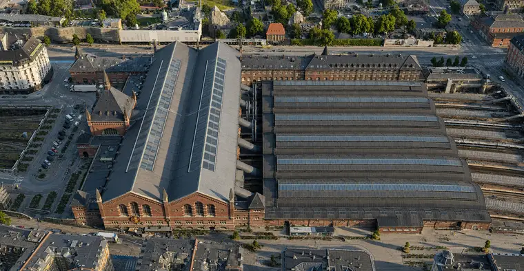 Aerial from the west: the great hall's double-ridged slate roof runs away from the camera behind its twin brick gable on Reventlowsgade, the spired entrance pavilion stands on the northern flank at the left, and the six platform halls fill the right half of the frame with the tracks running out beyond them.