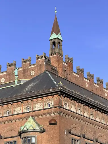 Billede af hjørne brystning med klokke tårnspir og rundeler set fra pladsen, i tegl, mørtel, natursten og skifer.