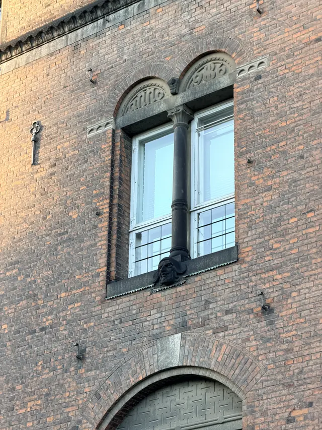 Billede af parret vindue med dato og udskåret hoved på gadefacaden, i tegl, natursten, træ og glas.