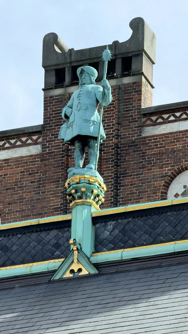 Billede af vægter med flettet våbenskjold på gadefacaden, i kobber, metal, tegl og mørtel.
