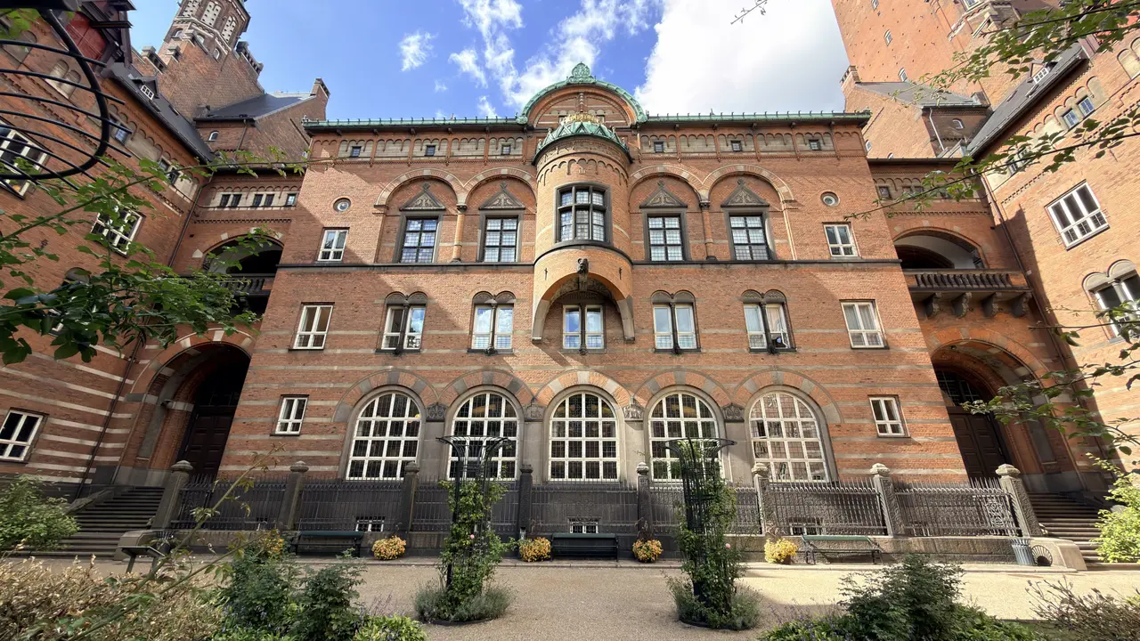 Gårdens havefacade set fra haven: fem høje rundbuede vinduer på en stribet sokkel af tegl og sten, en rund karnap med grønt kobbertag og forgyldt krone, og tårnet bagved mod blå himmel.