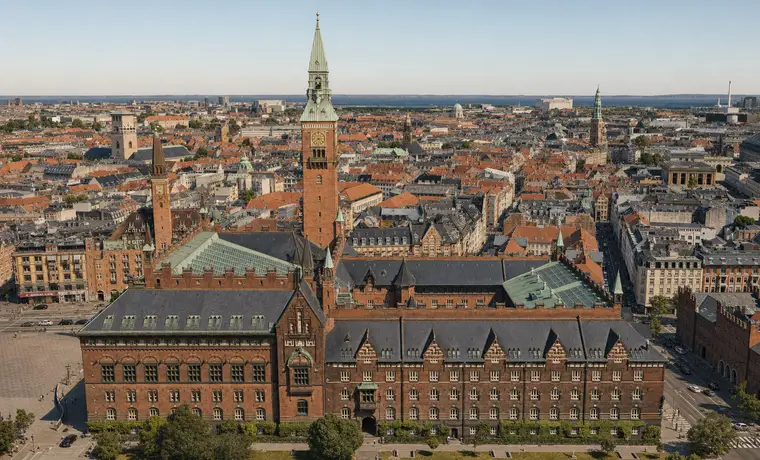 Københavns Rådhus set skråt fra luften med tårn, teglfacader, tagflader og Københavns tætte bymidte i baggrunden.