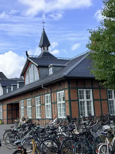 A long section of Østerport Station’s red-brick and green-painted timber street facade rises behind bicycle parking, with a lunette gable, slate roofs, central clock tower and spire partly framed by a tree.