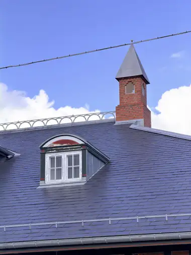 A red-brick chimney stack with a tall pyramidal metal cap rises above the slate roof beside a painted timber dormer.