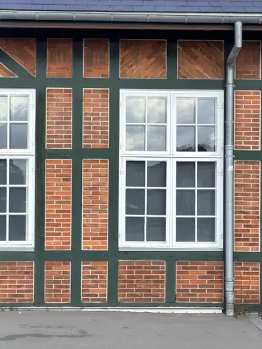 A close facade section shows green-painted timber framing, patterned red-brick infill, white multi-pane windows, slate roofing, and a zinc rainwater pipe.