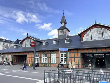 A wide view of the long street facade with patterned brick-and-timber framing, slate roofs, two lunette gables, and the central clock tower and spire.