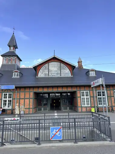 The street facade is shown with red brick infill, dark green timber framing, a large lunette window, slate roofs, and the clock tower and spire at left.