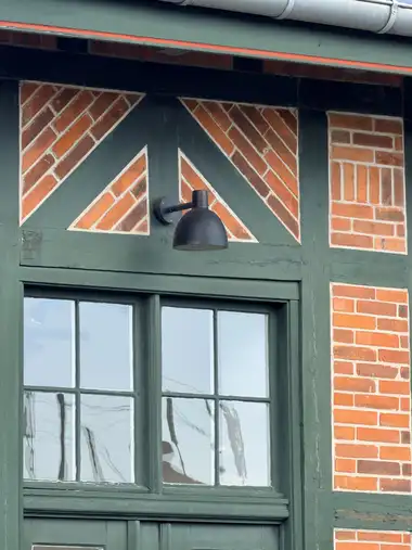 A green-painted timber-framed window bay with red brick infill laid in mirrored diagonal patterns, a wall lamp, and a zinc gutter above.