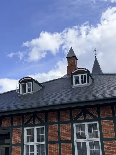 The steep slate roof is shown from below with two dormer windows, a brick chimney, a pointed roof turret and a weathervane above the half-timbered facade.