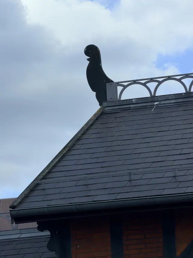 A flat wooden figure with a bird-like head at the end of a roof ridge, beside an iron cresting of small loops.