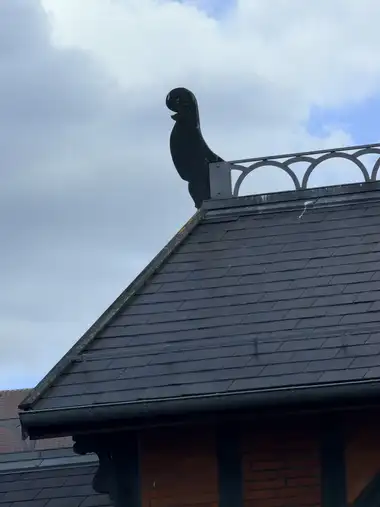 A flat animal-shaped finial stands at the apex of a slate-clad gable beside a decorative metal ridge railing.