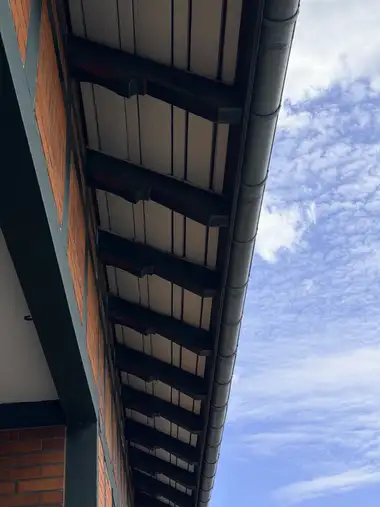 The frame looks upward along the deep boarded eaves, showing repeated dark-painted timber brackets with red accents, a metal gutter and part of the brick facade against the sky.