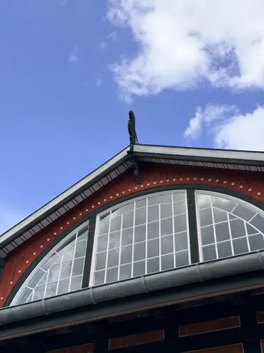 A low-angle view of the red-and-green timber gable with its large glazed lunette, metal gutter and flat cut-out ridge figure against the sky.