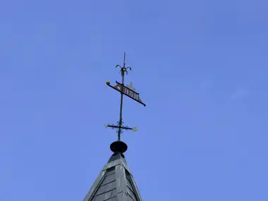 The station spire rises into a clear blue sky, topped by an ornate metal weathervane with a red DSB pennant, directional cross and decorative emblems.
