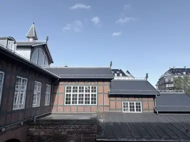 An upper trackside section of the station shows green-painted timber framing with patterned red-brick infill, large white-painted windows, adjoining slate and metal roofs, a lunette gable, a small spire, and silhouetted ridge-end figures.