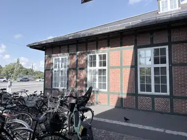 A street-side section of the station facade shows green-painted timber framing, patterned red-brick infill, white multi-pane windows, and a slate roof above parked bicycles.