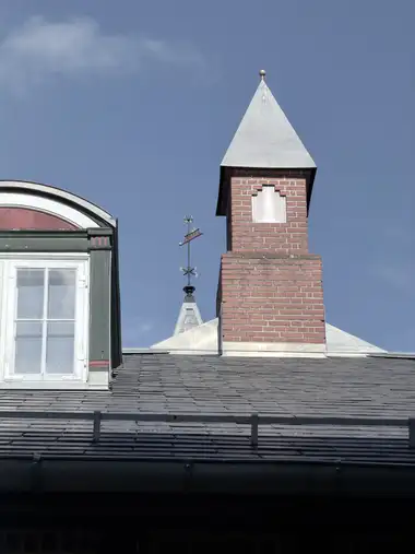 A tall red-brick chimney with a pyramidal metal cap rises above the slate roof, with a dormer and the station weathervane behind it.