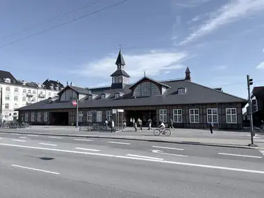 A wide frontal view shows the long, low station building with brick-and-timber walls, two lunette gables, a slate roof, and the central clock tower and spire.