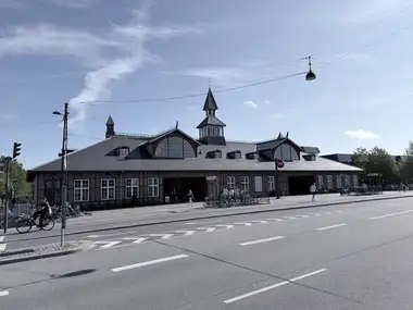 The complete long street facade of Østerport Station is shown across Oslo Plads, including its timber-framed brick walls, slate roof, twin lunette gables and central clock tower with spire.