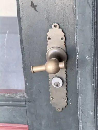 A close view of a brass lever handle, decorative backplate and modern cylinder lock mounted on a weathered green-painted glazed timber door.