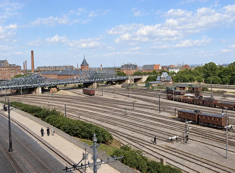 Elevated view across Østerport Station’s railway yard, with numerous tracks, rail carriages, a truss bridge, railway buildings and scattered pedestrians.