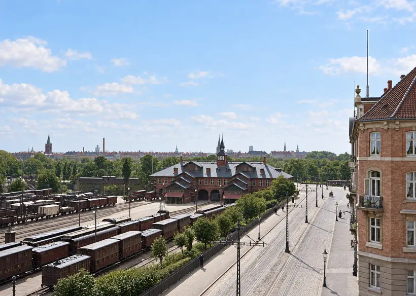 Elevated view of Østerport Station beside multiple railway tracks, parked carriages, a broad street, trees, and surrounding city buildings.