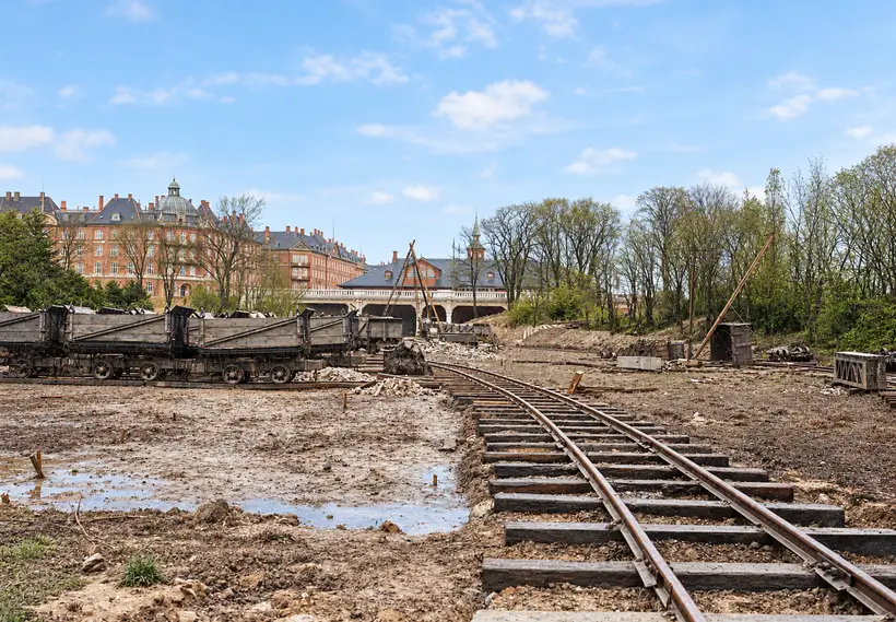 Rail tracks cross a cleared worksite toward Østerport Station, with open wagons, construction equipment, trees and large buildings behind.