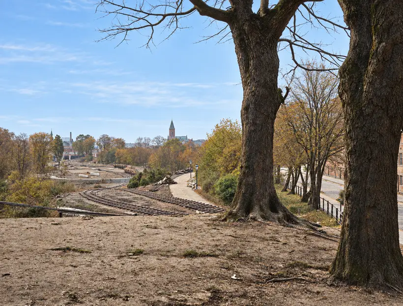Curving railway tracks cross an excavated landscape beside mature trees, with distant buildings and a church tower beyond.