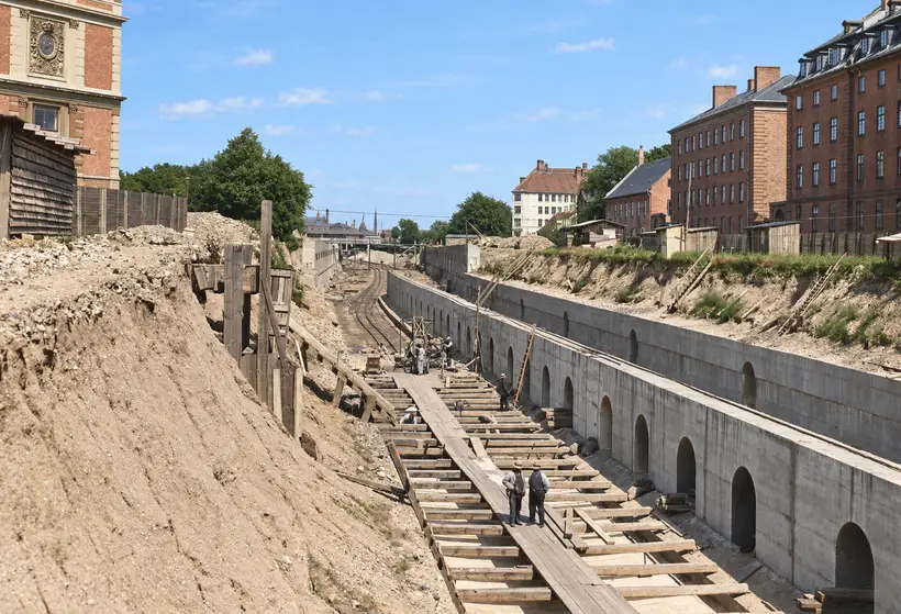 Workers stand on timber walkways beside railway tracks in a deep cutting bordered by concrete retaining walls and urban buildings.