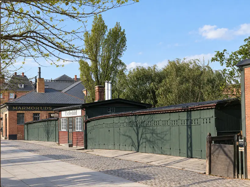Low station outbuildings and timber sheds line a cobbled lane beneath trees, with signs for bicycle storage and toilets.