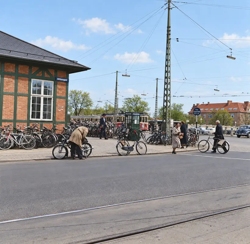 Street view with a brick-and-timber station building, parked bicycles, pedestrians, public transport, overhead wires, and tram tracks.