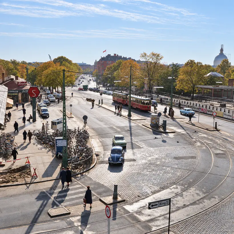 Elevated view across Oslo Plads with trams, cars, cyclists, pedestrians, overhead wires, station platforms and surrounding buildings.