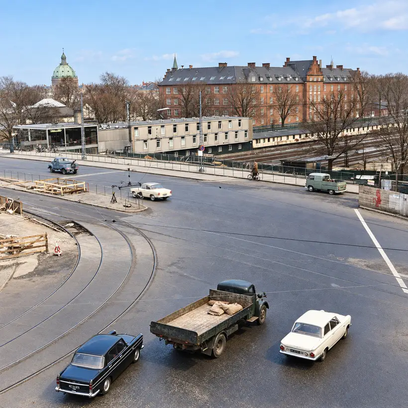 Elevated view across Oslo Plads traffic and tram tracks toward railway lines, low buildings, trees, and a large brick building.
