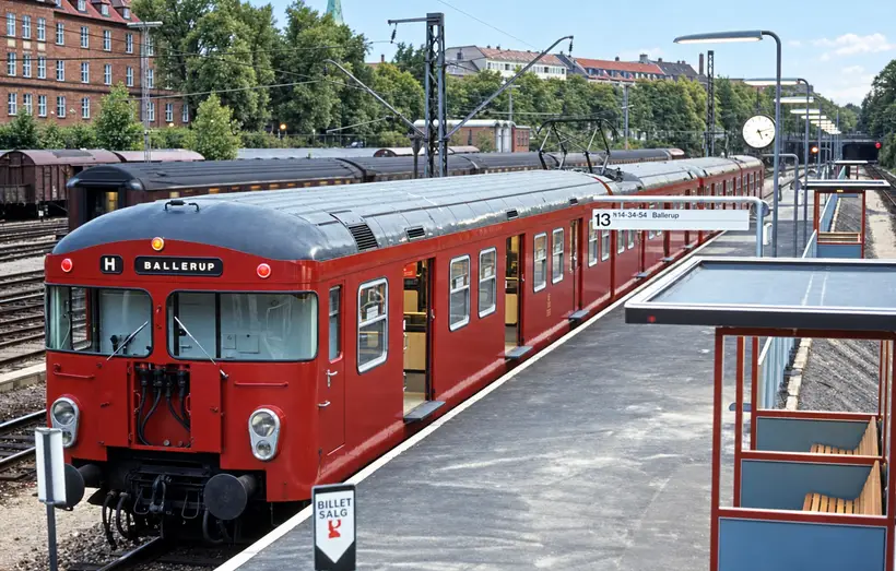 A DSB train stands beside an Østerport platform beneath overhead wires, with tracks, shelters, a clock, trees and buildings beyond.