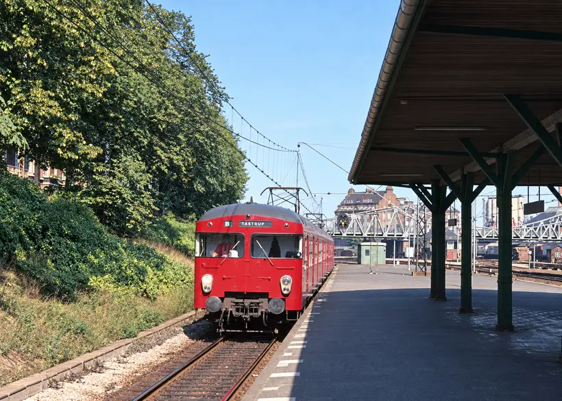 A passenger train approaches an open platform beneath a timber canopy, with overhead wires, tracks, trees and a footbridge beyond.