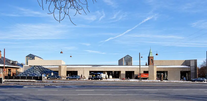 Street-facing overview of Østerport Station, with a long entrance façade, projecting skylights, pedestrians, bicycles, vehicles, snow and overhead wires.