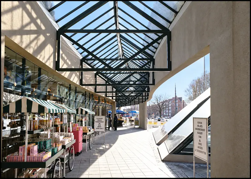 A covered station passage with exposed roof trusses, shop windows, bakery displays, pedestrians and stairs descending beside a curved wall.