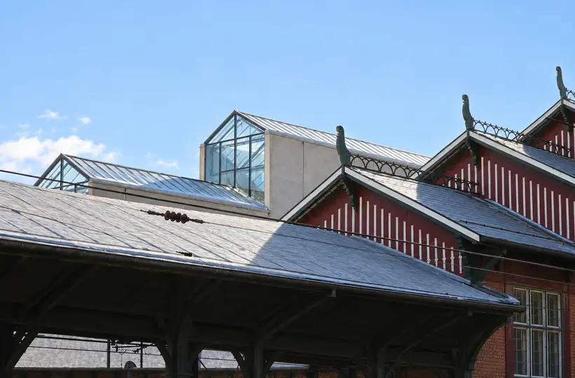 Glazed shopping-centre roofs rise behind the old station’s pitched roofs and ornamental gables, crossed by overhead railway wires.