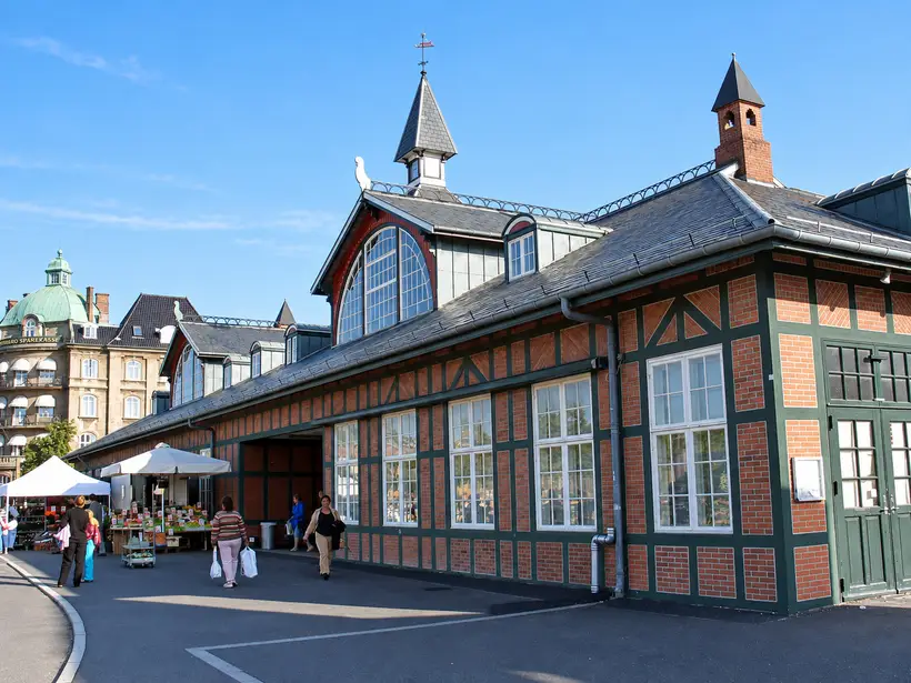 The station’s long framed façade and steep roof stand beside market stalls and pedestrians under a clear sky.