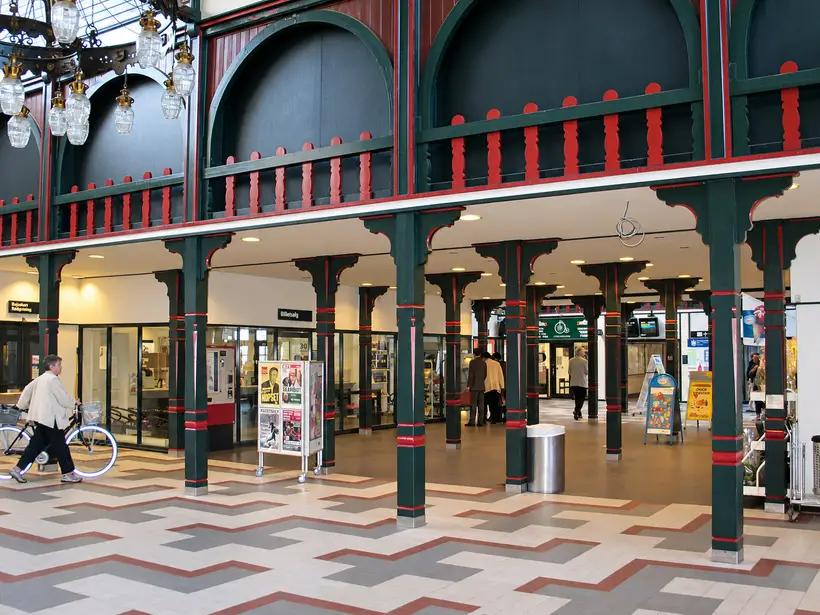 Station ticket hall with slender columns, arched upper panels, hanging lamps, ticket counters, bicycles, signs and passengers.