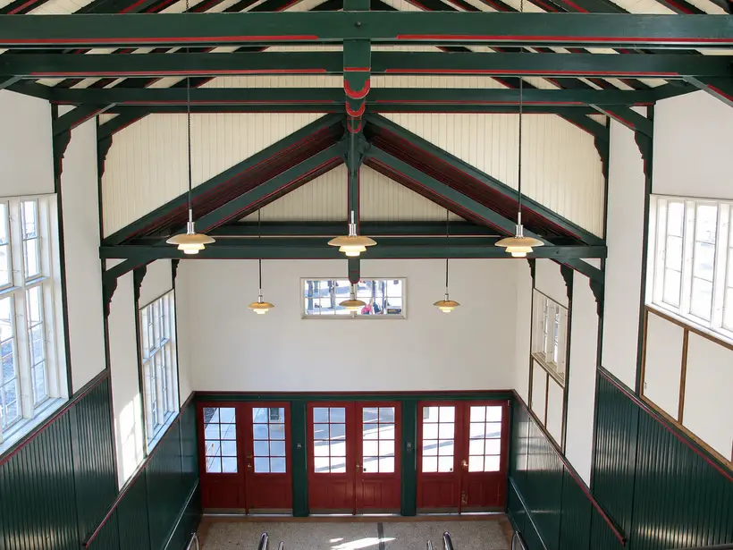Elevated interior view of a station hall with exposed timber roof trusses, suspended lamps, tall windows and glazed entrance doors.