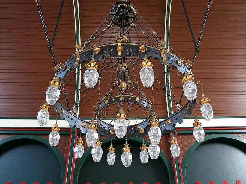 An elaborate circular chandelier with multiple caged glass lamps hangs beneath a panelled ceiling above arched wall sections.
