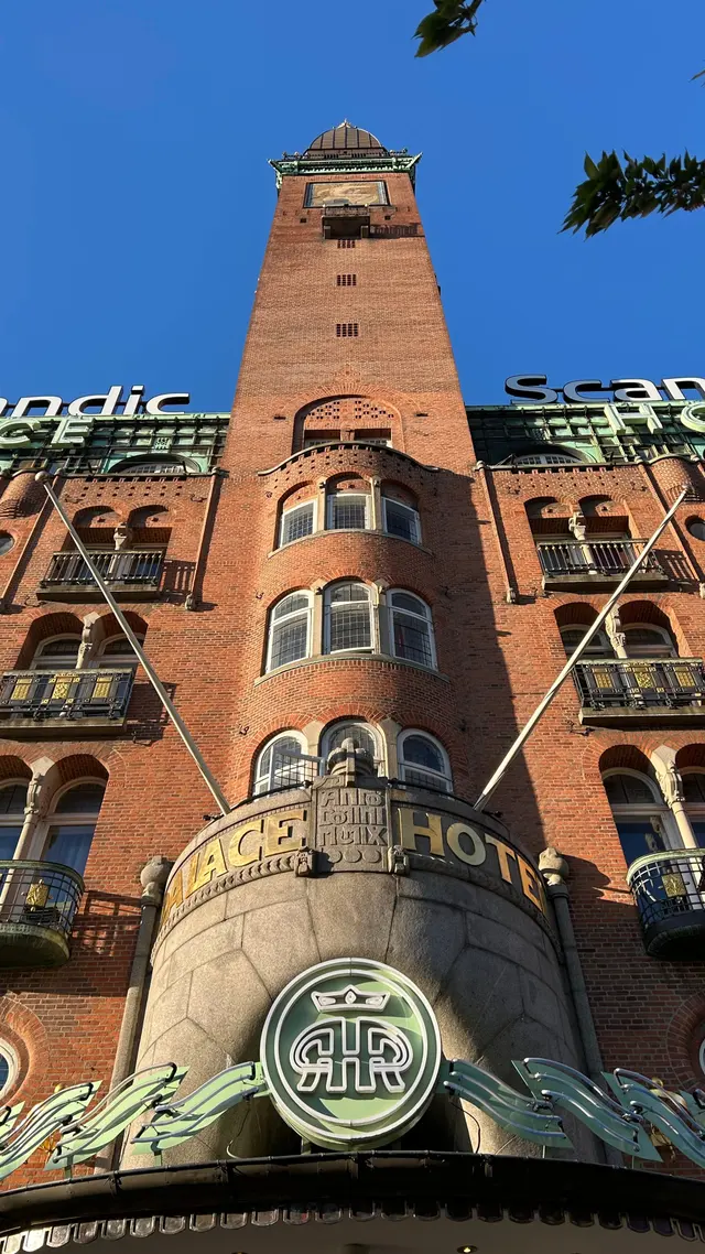 Set stejlt opad: tårnet med sit mosaikfelt og kobberkuplen, og forneden den buede granitbaldakin med PALACE og HOTEL i guld, ANNO DOMINI MCMIX hugget imellem dem, og det kronede monogram som lysskilt under.