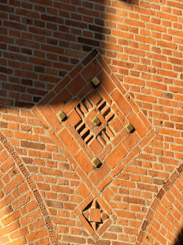 Blank mur i rødt tegl med et rudeformet felt af formsten og forgyldte klodser, og skråtstillede bånd af formsten, der hver bærer det samme lille mærke i en rektangulær ramme.