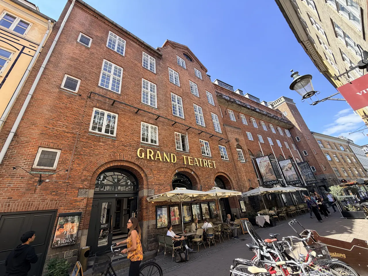 GRAND TEATRET i forgyldte bogstaver på den røde mur over biografens indgang, med caféborde og plakater i gadeplan.