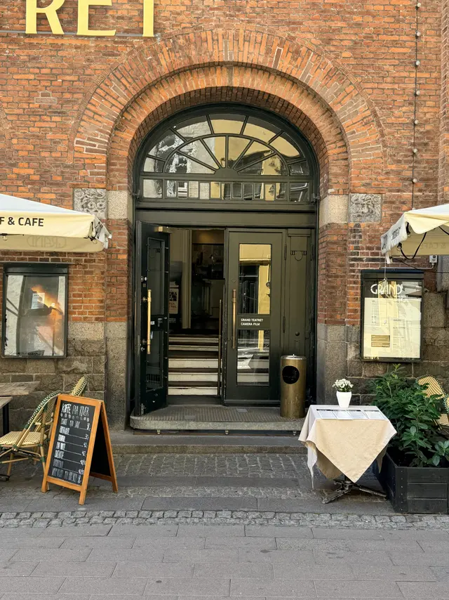 Indgangen til Grand Teatret i baggaden: en bred murbue over mørkegrønt træværk, hvor én dør står åben over en trappe af lyse trin.