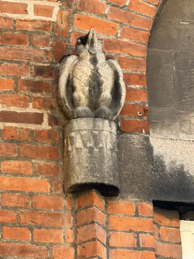 En hugget fugl med foldede vinger på en lille cylindrisk konsol, hvor vinduesbuen sætter an.