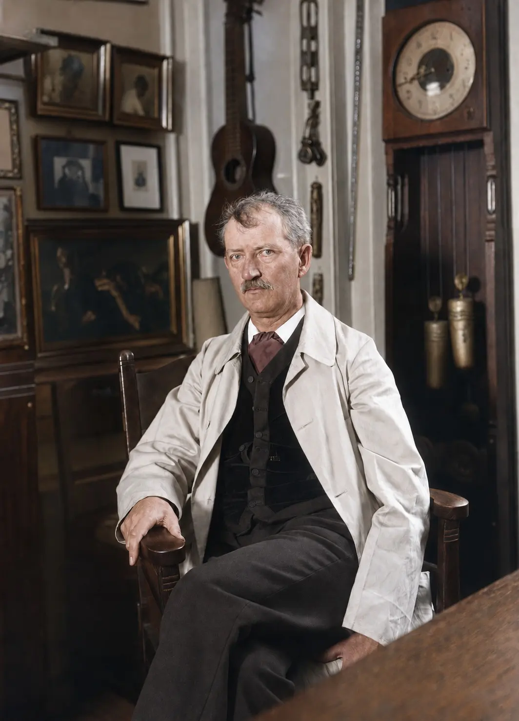 Anton Rosen sidder i en armstol i lys arbejdskittel over mørk vest og halsbind, med indrammede billeder, en guitar og et standur bag sig.
